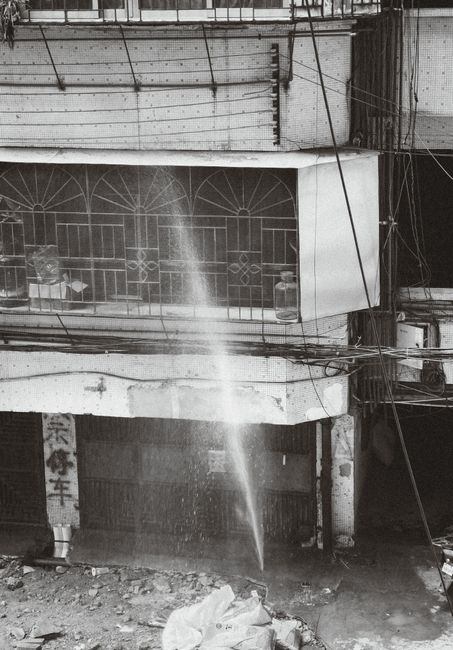 The image depicts a street scene in Black and White, showcasing an unfinished or under-construction building façade with a decorative iron grille or window screen, partially obscured by dust and debr