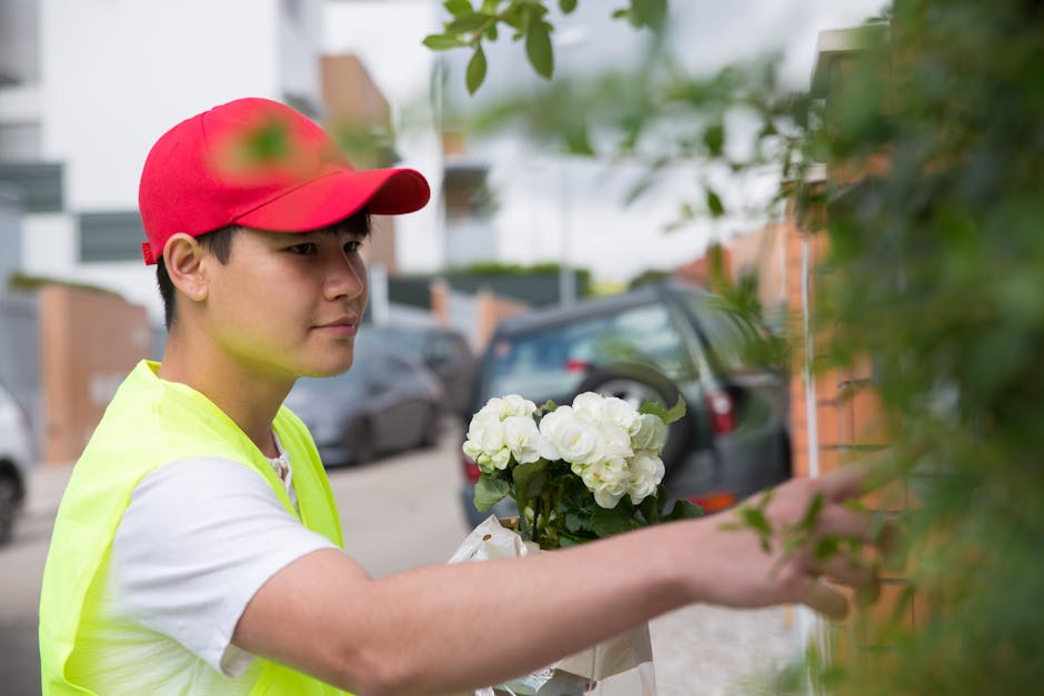 A young male flower delivery courier wearing a red cap and a high-visibility yellow vest is seen placing a bouquet of fresh white hydrangeas wrapped in clear cellophane against a green hedge outside a