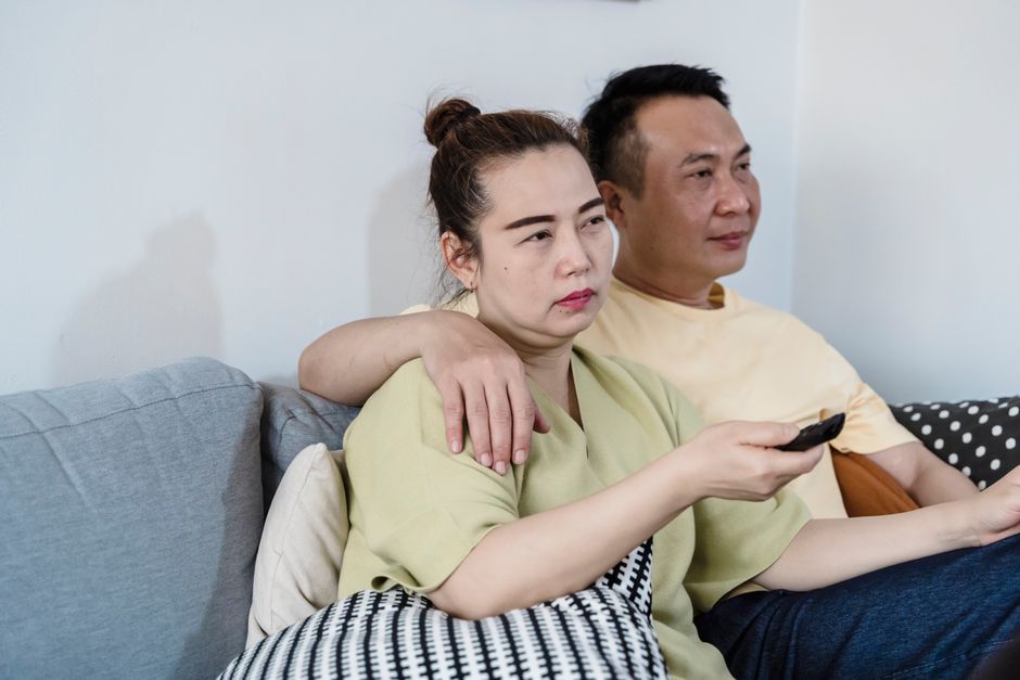 A woman with dark hair tied in a bun, wearing a light green top, is sitting on a grey sofa, holding a remote control in her right hand. She appears somewhat disinterested or annoyed. Behind her, a man