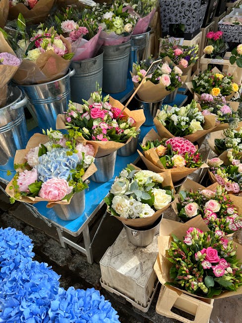 A vibrant display of fresh flower bouquets arranged in galvanized metal buckets and wrapped in brown paper, featuring a variety of pastel pink, white, and lavender roses, hydrangeas, and other blooms.