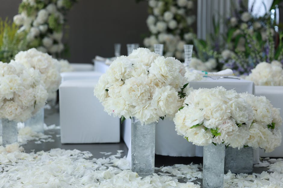 A floral arrangement featuring large, round bouquets of white carnations and peonies, arranged in tall, clear glass and concrete vases placed on a table covered with white flower petals. The flowers a