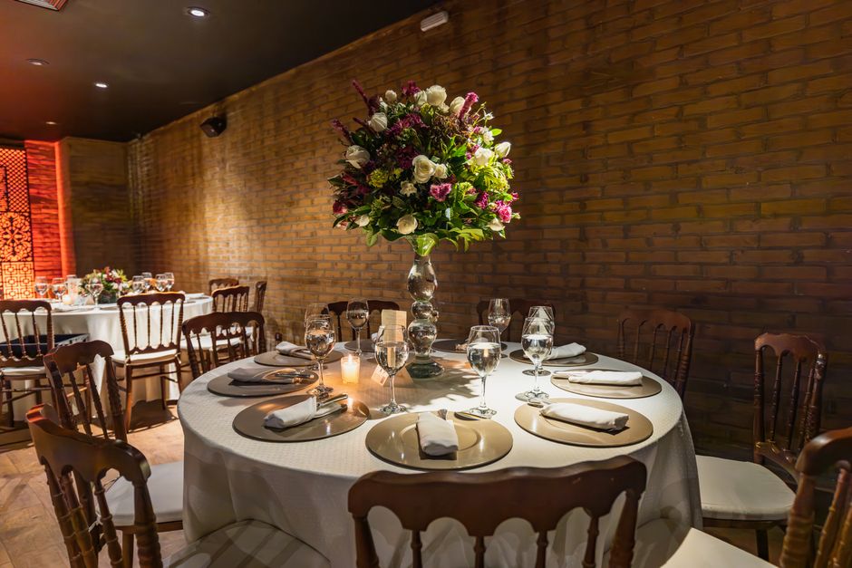 A round table set for an event features a tall, elegant floral arrangement with white roses, purple and pink flowers, and lush green foliage in a clear glass vase. The table is draped with a white tab